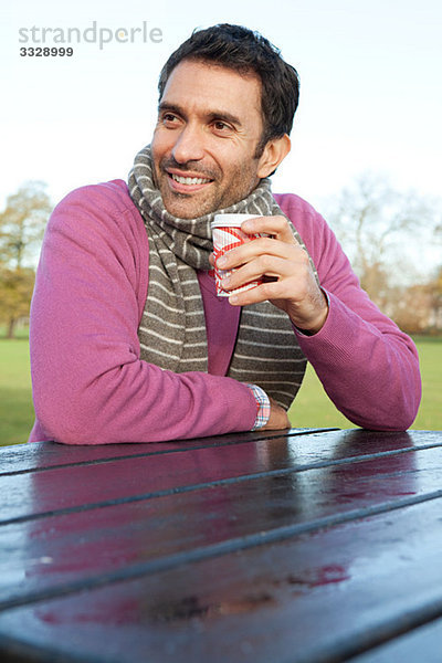 Ein Mann  der an einem Parktisch sitzt und ein heißes Getränk hält.