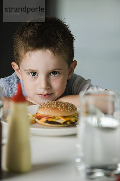 Kleiner Junge am Tisch sitzend mit Cheeseburger auf Teller  Kopf auf Armen liegend