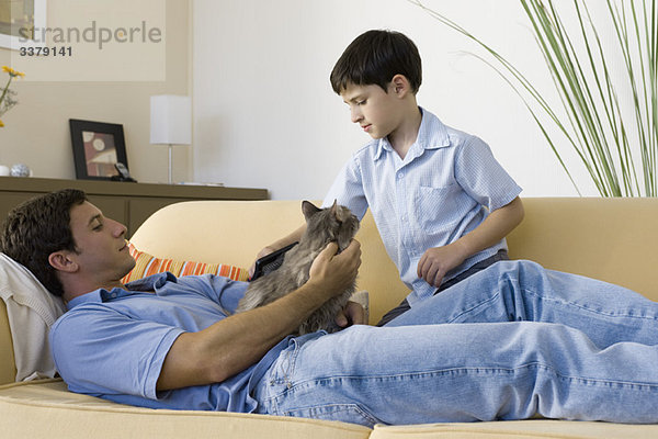 Mann entspannt mit Katze auf dem Schoß  junger Sohn bürstet Katze