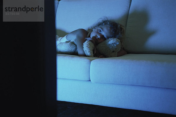 Mädchen auf dem Sofa liegend mit Teddybär  fernsehend