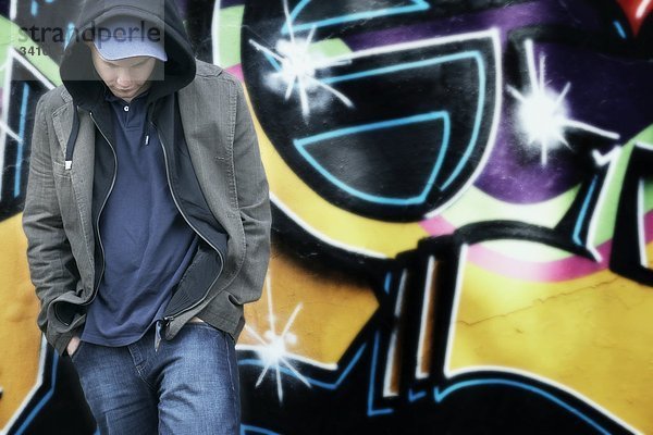 Junger Mann mit Kapuzenjacke und Kappe vor einer Graffitiwand