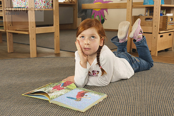 Deutschland  Mädchen (6-7) im Kinderzimmer auf Teppich liegend beim Lesen eines Buches