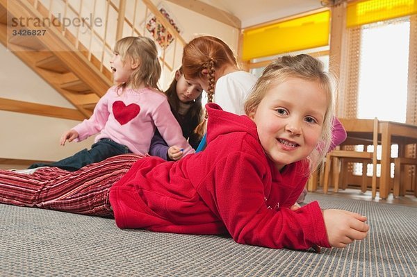 Deutschland  Kinder im Kindergarten  Mädchen (6-7) auf dem Boden liegend  Kinder im Hintergrund zusammen spielend