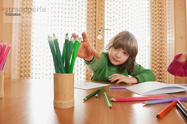 Deutschland  Junge (6-7) im Kinderzimmer am Tisch sitzend  nach Buntstift greifend  Portrait