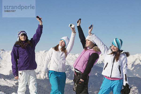 Vier Teenagermädchen in Skikleidung  mit ihren Handys in der Luft.