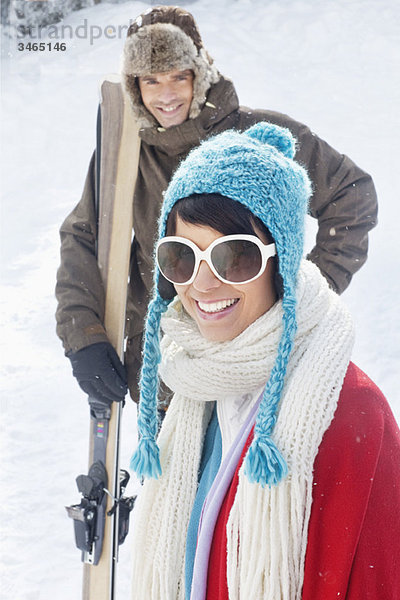 Junge Frau in Winterkleidung lächelnd vor der Kamera  Mann mit Skiern im Hintergrund