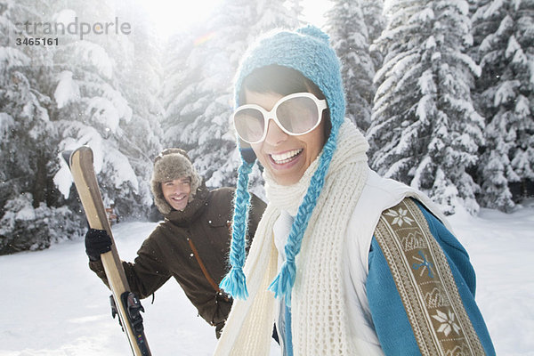 Junge Frau in Winterkleidung lächelnd vor der Kamera  Mann mit Skiern im Hintergrund