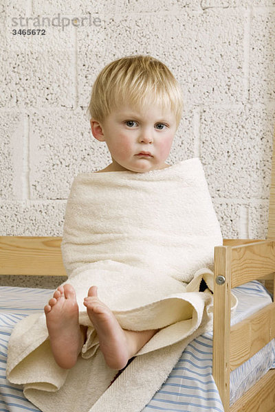 Ein unglücklicher kleiner Junge in ein Handtuch gewickelt  das auf einem Bett sitzt.
