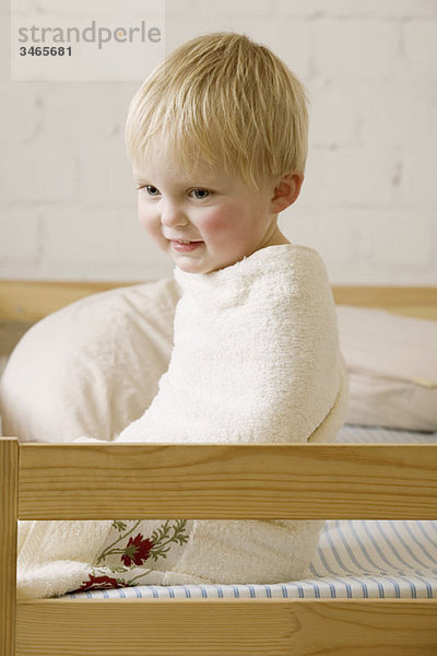 Ein fröhlicher kleiner Junge in ein Handtuch gewickelt  das auf einem Bett sitzt.