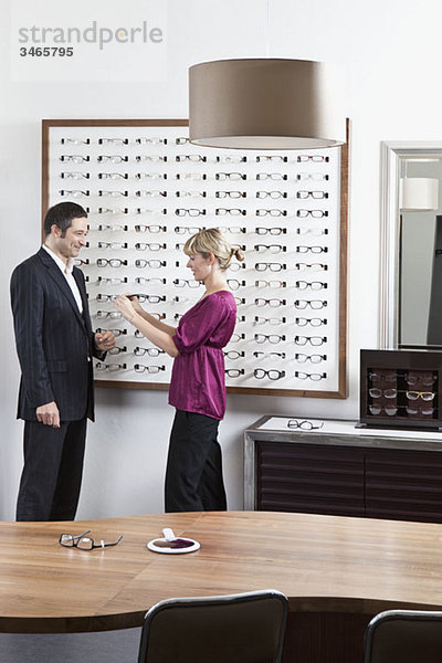 Ein Optiker  der einem Mann in einem Brillengeschäft eine Brille vorhält.