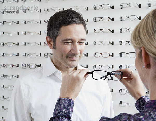 Ein Optiker  der einem Mann in einem Brillengeschäft eine Brille vorhält.