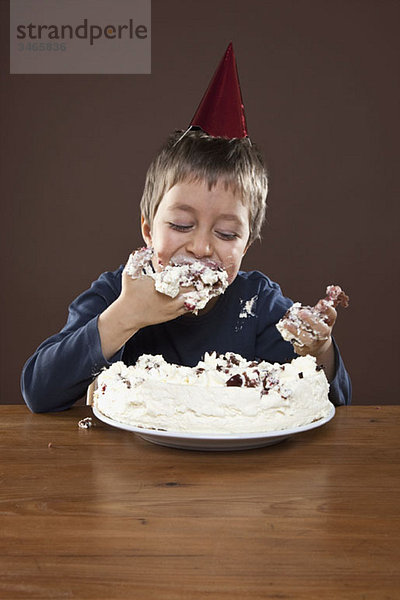 Ein Junge mit einem Partyhut  der Geburtstagskuchen mit den Händen isst  Atelieraufnahme