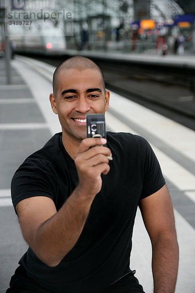 Mann sitzt auf einem Bahnsteig und macht ein Foto mit seinem Handy