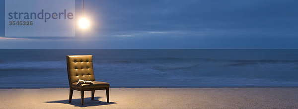 Glühbirne beleuchtet über Stuhl mit offenem Buch am Strand bei Nacht