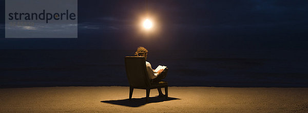 Mann sitzt nachts im Stuhl unter Glühbirne am Strand  liest Buch