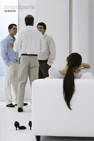 Frau entspannt im Sessel  während männliche Kollegen im Hintergrund plaudern