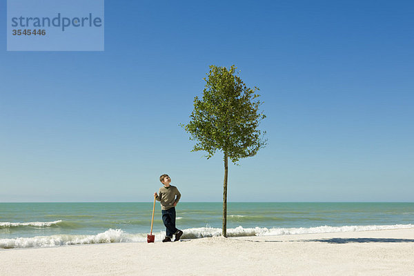 Junge stehend mit Schaufel neben Baum am Strand gepflanzt