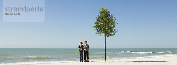 Kinder stehen mit Schaufeln neben einem am Strand gepflanzten Baum.