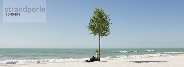 Junge liest Buch unter einem Baum am Strand