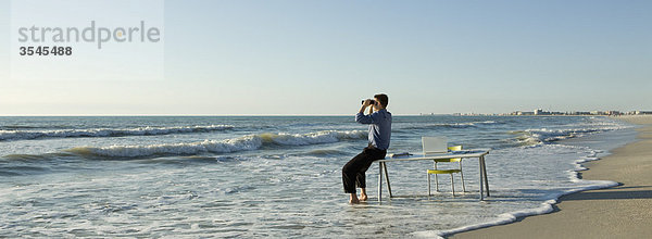Geschäftsmann sitzt auf dem Schreibtisch am Strand und schaut durch ein Fernglas auf See.