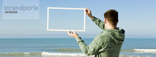Möwen  die über dem Meer fliegen  gerahmt in einem Bilderrahmen  der von einem Mann am Strand hochgehalten wird.