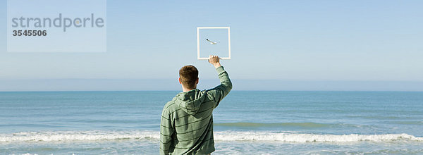 Mann am Strand hält Bilderrahmen hoch und fängt das Bild einer Möwe ein  die gegen den blauen Himmel fliegt.