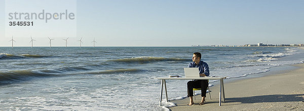 Mann arbeitet am Schreibtisch am Strand  Windkraftanlagen am Horizont