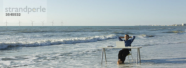 Mann entspannt am Schreibtisch am Strand  Windkraftanlagen am Horizont