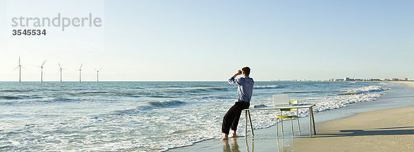 Mann auf Schreibtischkante am Wasserrand am Strand mit Blick auf Offshore-Windkraftanlagen am Horizont