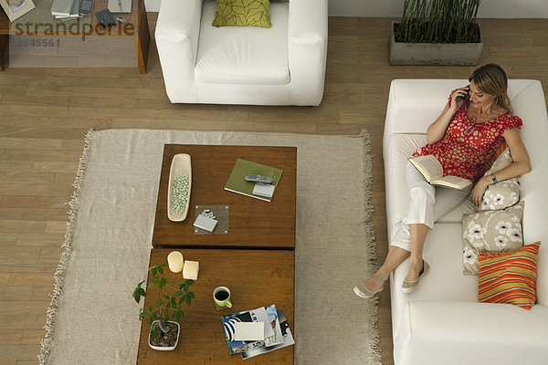 Frau entspannt auf dem Sofa mit Buch auf dem Schoß  spricht auf dem Handy