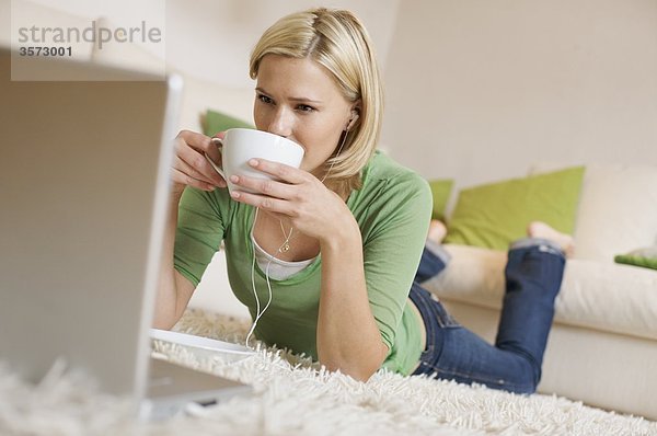 Junge Frau liegt auf einem Teppich mit Kaffeetasse  Ohrstöpseln und Laptop