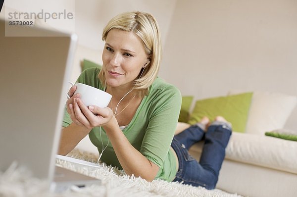 Junge Frau liegt auf einem Teppich mit Kaffeetasse  Ohrstöpseln und Laptop