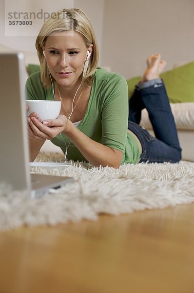 Junge Frau liegt auf einem Teppich mit Kaffeetasse  Ohrstöpseln und Laptop