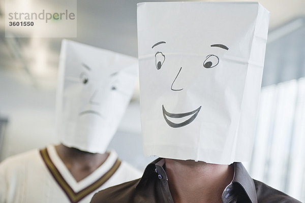 Zwei Geschäftsleute  die Papiertüten mit fröhlichen und traurigen Gesichtern tragen.