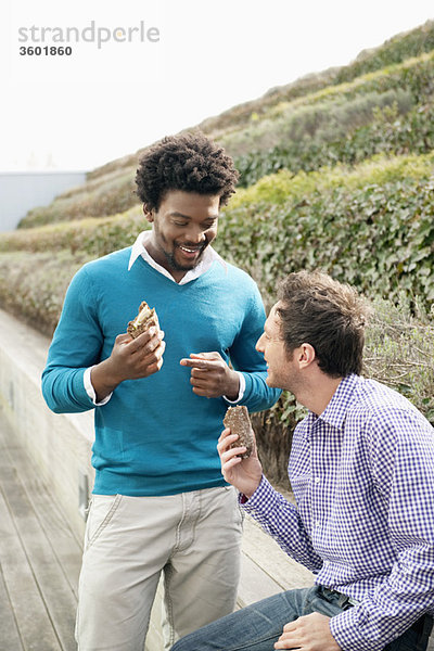 Zwei Freunde essen Sandwiches auf einer Promenade.