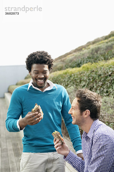 Zwei Freunde essen Sandwiches auf einer Promenade.
