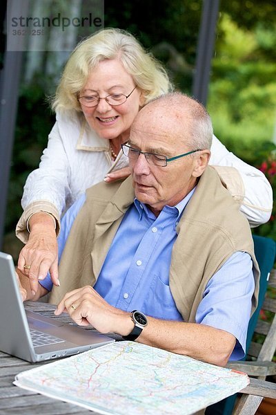 Seniorenpaar auf der Terrasse mit Laptop und Straßenkarte