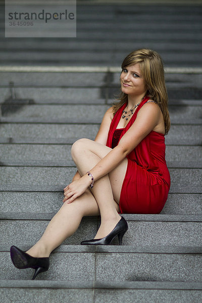 Junge Frau in rotem Kleid und hochhackigen schwarzen Schuhen sitzt auf Steintreppe