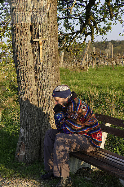 Mann auf Bank unter Baum mit Kreuz bei Großau bei Bad Vöslau Niederösterreich Österreich