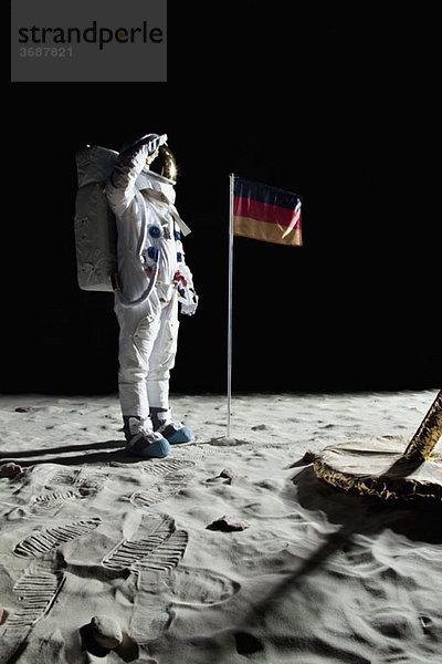 Ein Astronaut  der in der Nähe einer Mondlandefähre steht  grüßt eine deutsche Flagge.