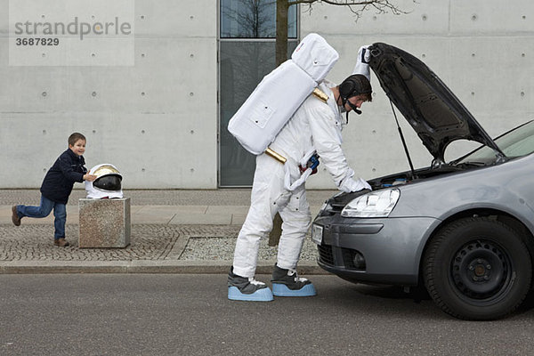 Ein Kind stiehlt einen Weltraumhelm  während ein Astronaut sein kaputtes Auto repariert.