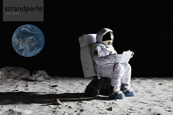 Ein Astronaut auf dem Mond  der mit einem Laptop auf einem Felsen sitzt.