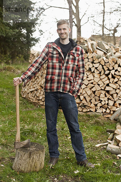 Ein Mann  der eine Axt hält und vor einem Holzhaufen steht.