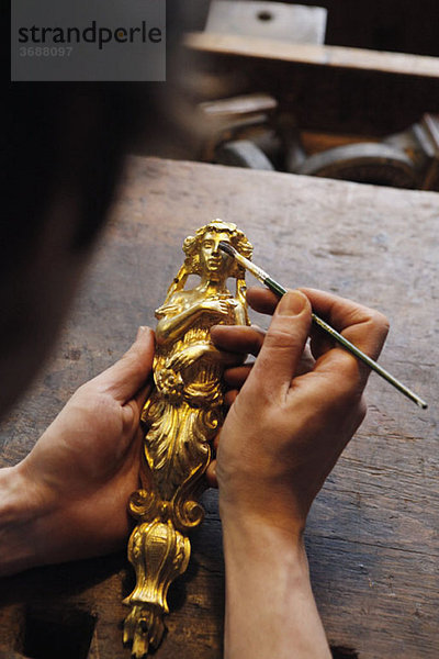 Detail einer Person  die eine vergoldete Figur und einen Pinsel hält.