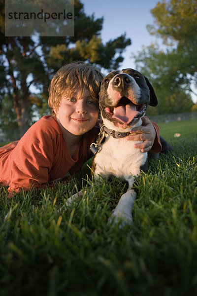 Porträt eines Jungen  der mit einem Hund auf Gras liegt.