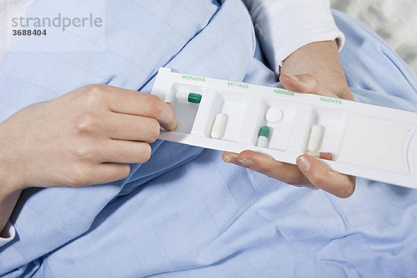 Ein Patient in einem Krankenhausbett  der eine Pille von einem Pillenorganizer nimmt  konzentriert sich auf die Hände.