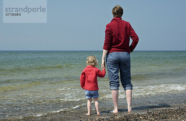 Junge Frau steht mit ihrer kleinen Tochter am Strand und schaut aufs Meer