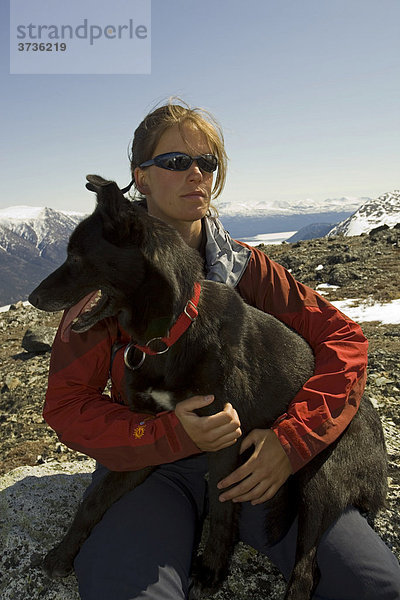 Wanderin rastet auf einem Fels  junge Frau mit Schlittenhund  Alaskan Husky  dahinter Berge  Kusawa Gebirgskette  Yukon Territory  Kanada  Nordamerika