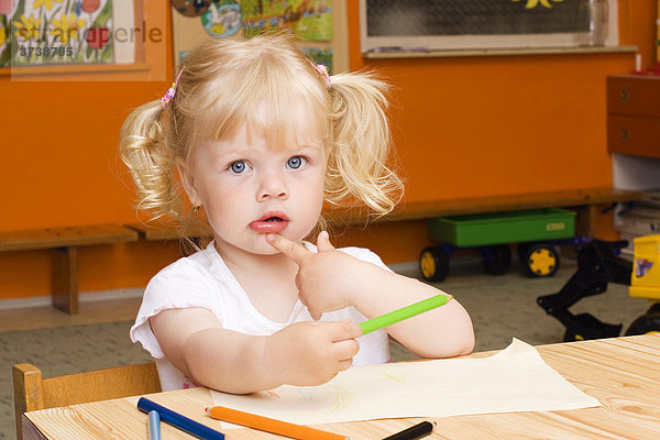 Blondes kleines Mädchen  3 Jahre alt  sitzt mit Kreiden an einem Tisch