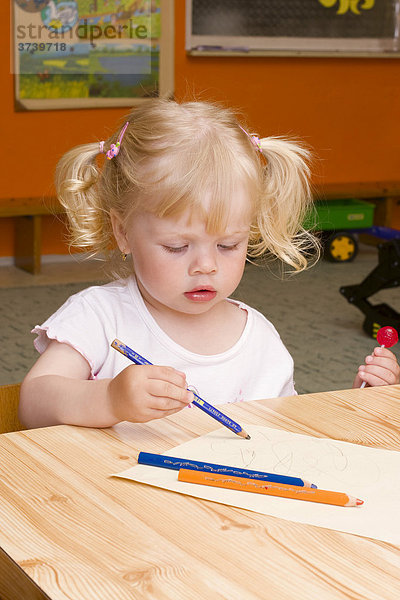 Zweijähriges Mädchen sitzt mit Buntstiften am Tisch